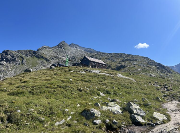 Eine Berghütte mit einer grünen Fahne steht an einem grasbewachsenen Hang unter einem strahlend blauen Himmel und felsigen Gipfeln.