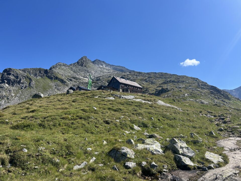 Eine Berghütte mit einer grünen Fahne steht an einem grasbewachsenen Hang unter einem strahlend blauen Himmel und felsigen Gipfeln.