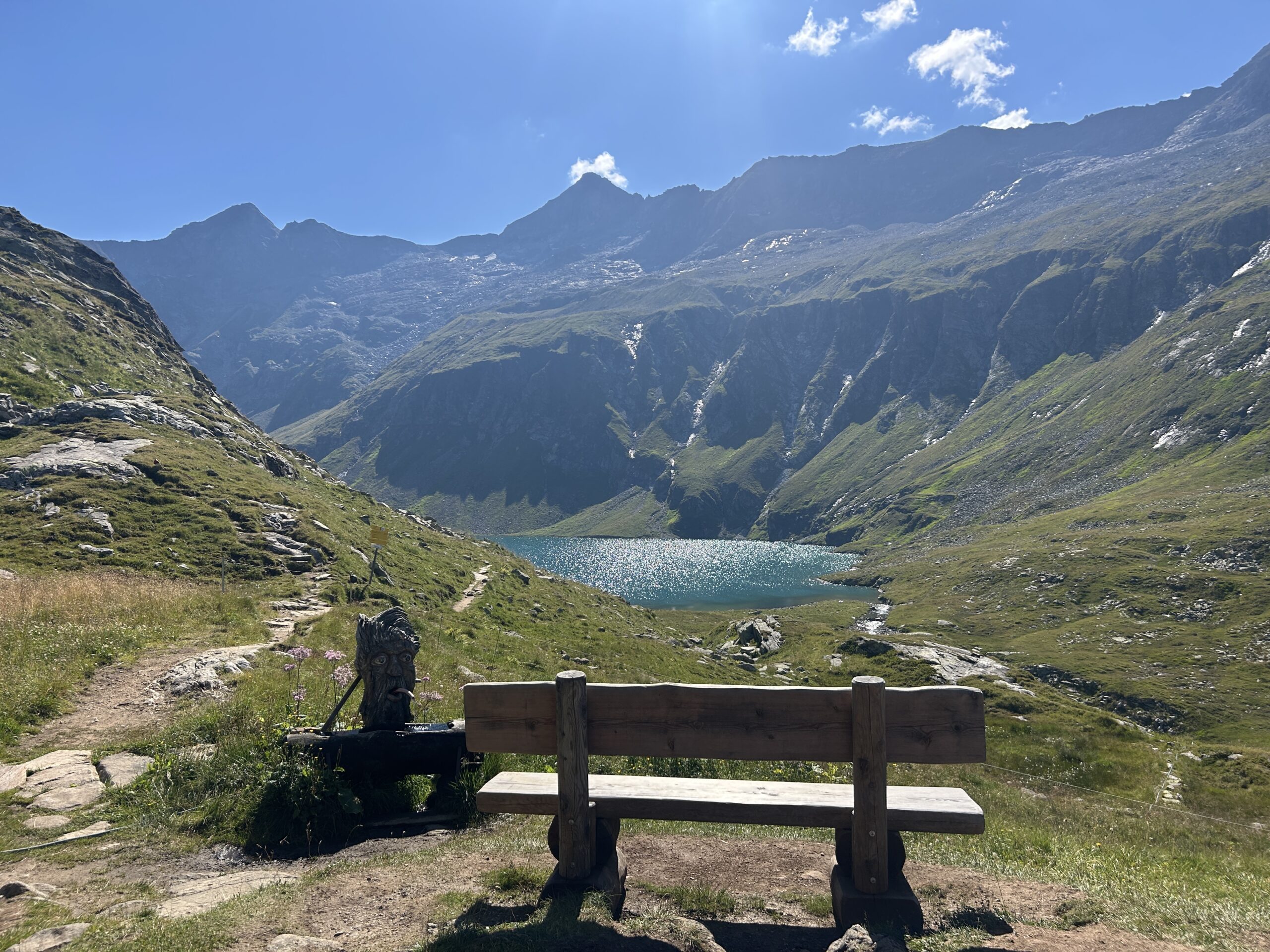 Von einer Holzbank aus hat man einen Blick auf einen Bergsee mit sonnenbeschienenen, gr&uuml;nen H&uuml;geln und felsigen Gipfeln unter blauem Himmel.