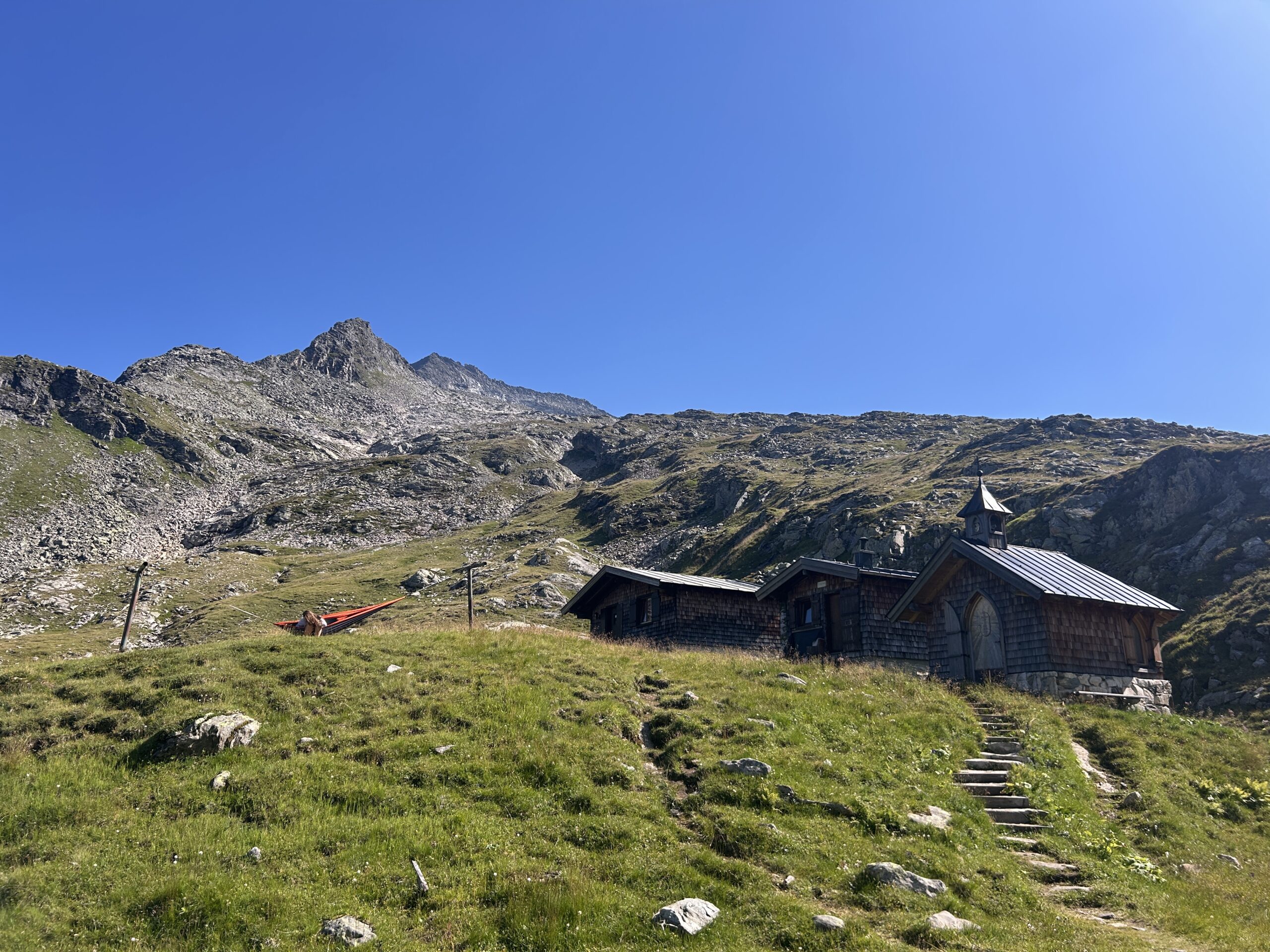 Eine kleine Holzh&uuml;tte und eine Kapelle stehen auf einem grasbewachsenen Hang, im Hintergrund ragen felsige Berge empor, &uuml;ber ihnen erstreckt sich ein klarer Himmel.