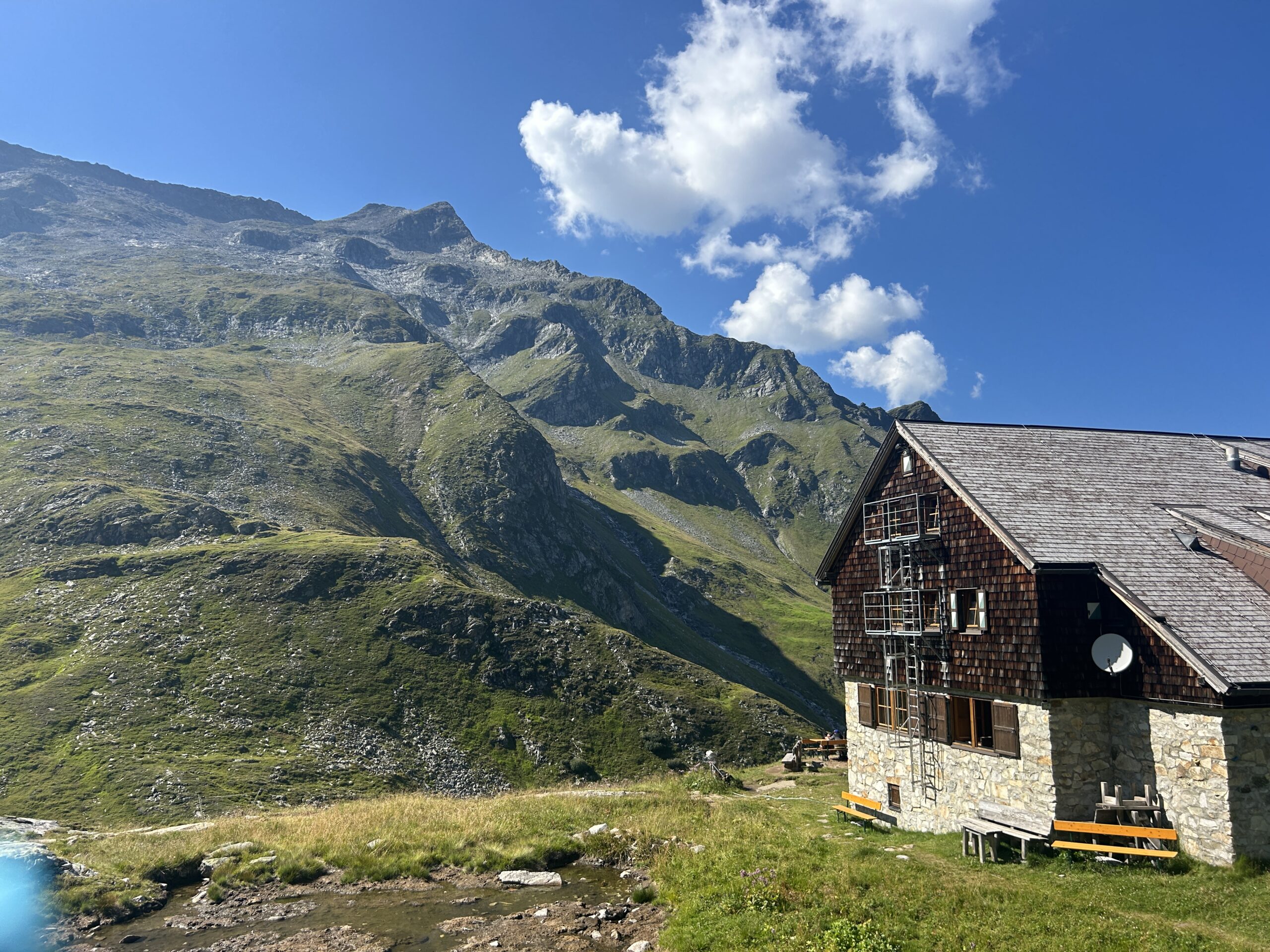 Eine Steinh&uuml;tte in der N&auml;he eines kleinen Teiches steht unter einem bewachsenen Berghang vor einem blauen Himmel mit Wolken.
