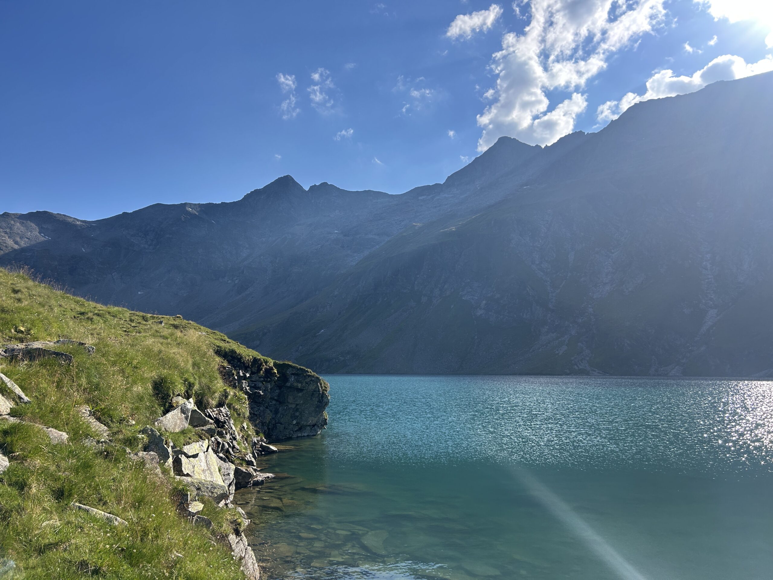 Ein klarer See mit türkisfarbenem Wasser, grasbewachsenem Ufer, felsigen Bergen im Hintergrund und blauem Himmel mit vereinzelten Wolken.