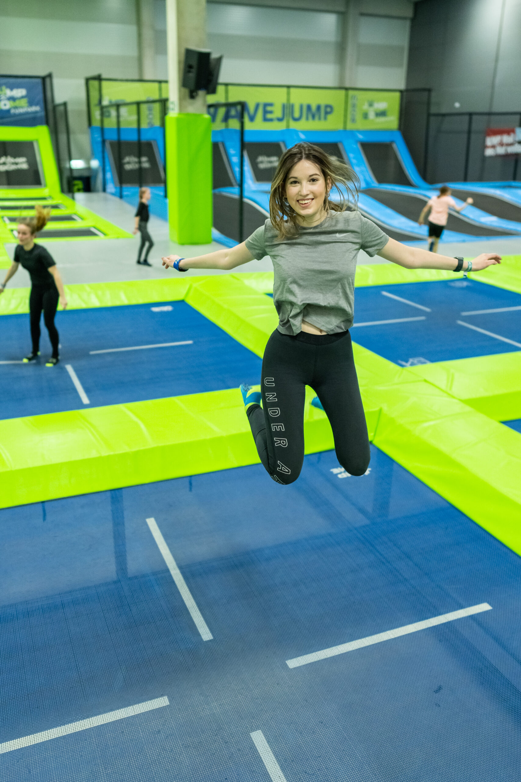 Eine junge Frau springt l&auml;chelnd auf einem Trampolin in einem Raum, w&auml;hrend im Hintergrund andere ebenfalls springen.
