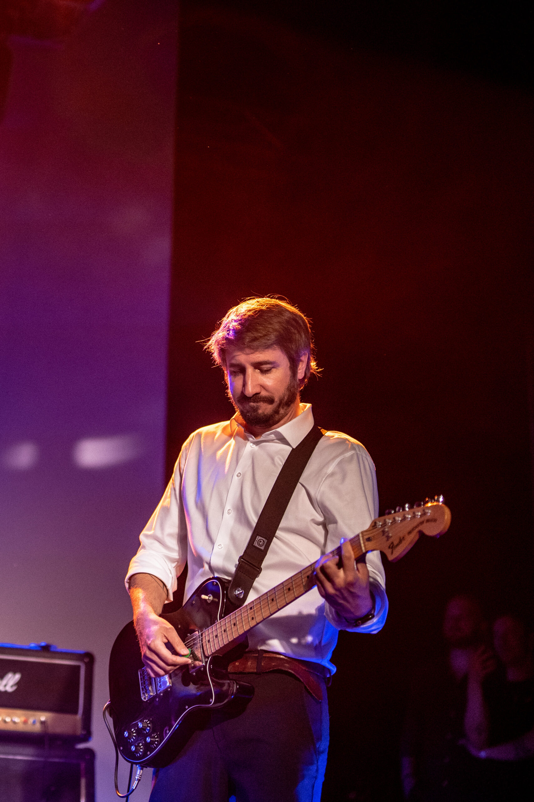 Ein Mann in einem wei&szlig;en Hemd, der auf einer B&uuml;hne bei warmer Beleuchtung E-Gitarre spielt.