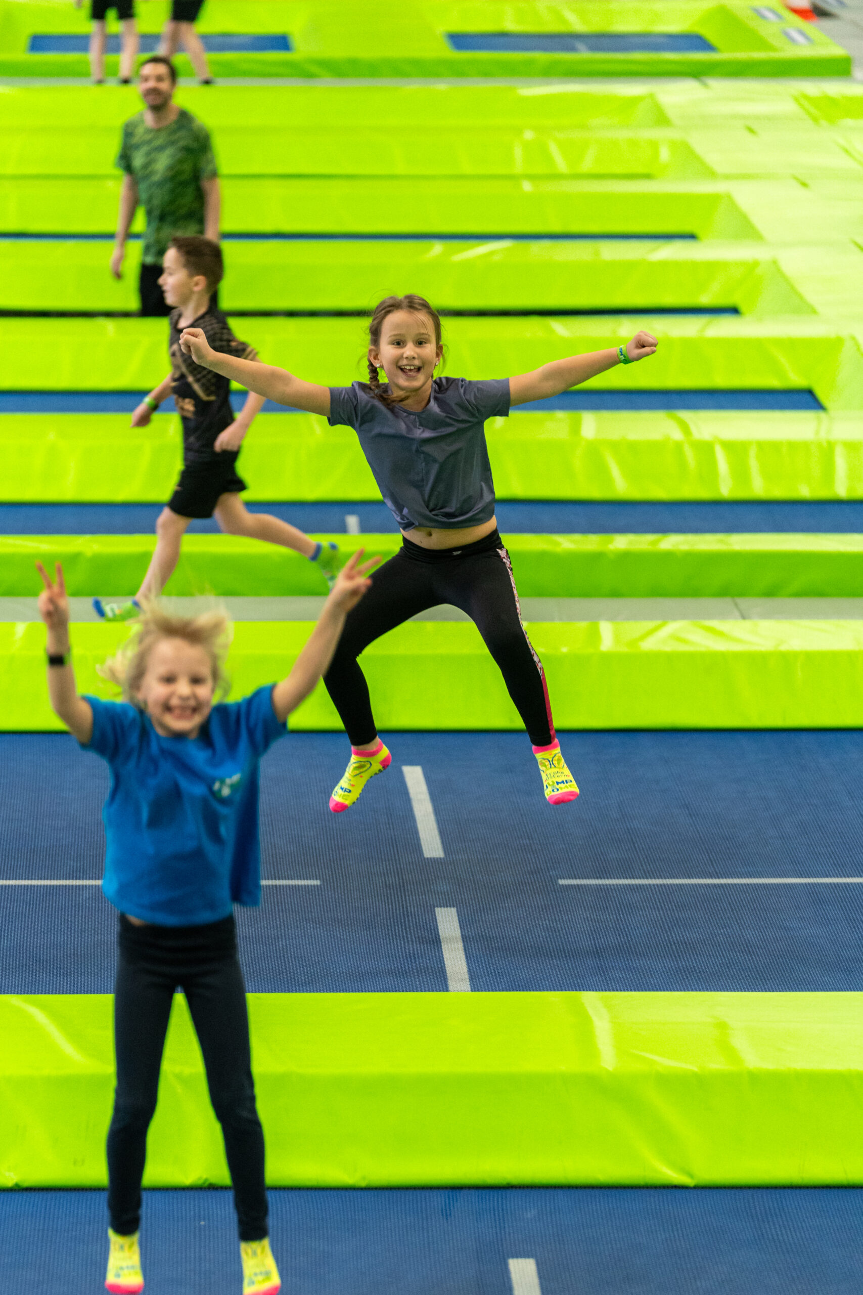 Zwei Kinder, die auf leuchtend gr&uuml;nen Trampolinen springen und l&auml;cheln, im Hintergrund sind weitere Personen zu sehen.