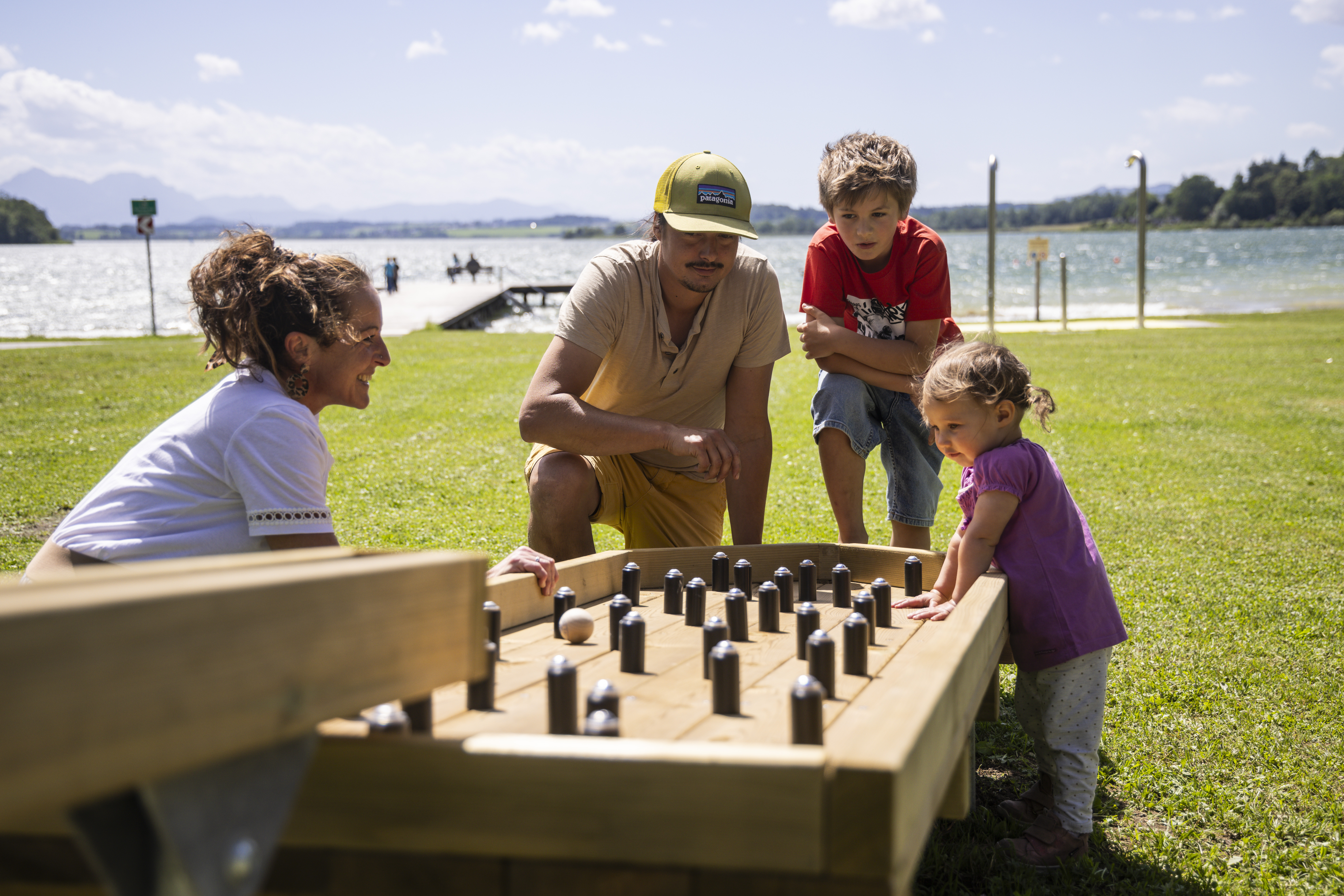 Vier Personen spielen bei sonnigem Wetter auf einer Wiese in der Nähe eines Gewässers ein Brettspiel aus Holz.
