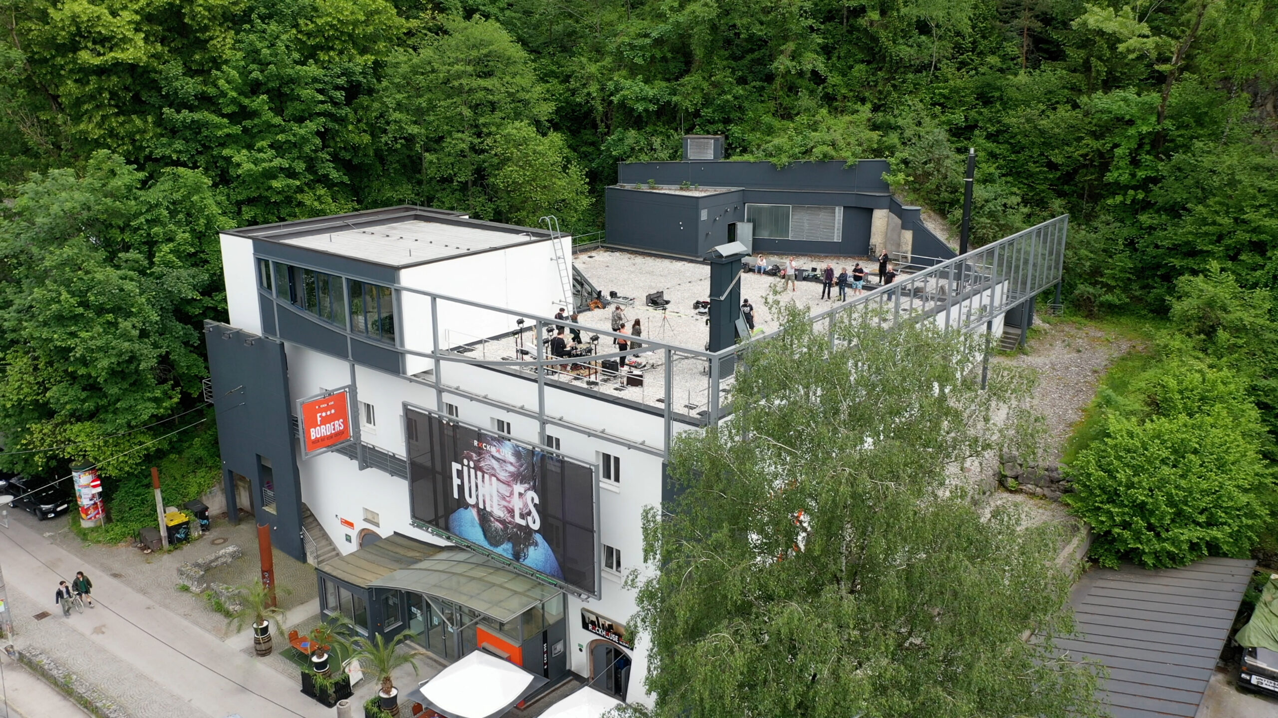 Luftaufnahme eines wei&szlig;en Geb&auml;udes mit Sitzgelegenheiten auf dem Dach, umgeben von B&auml;umen und Gr&uuml;nfl&auml;chen.