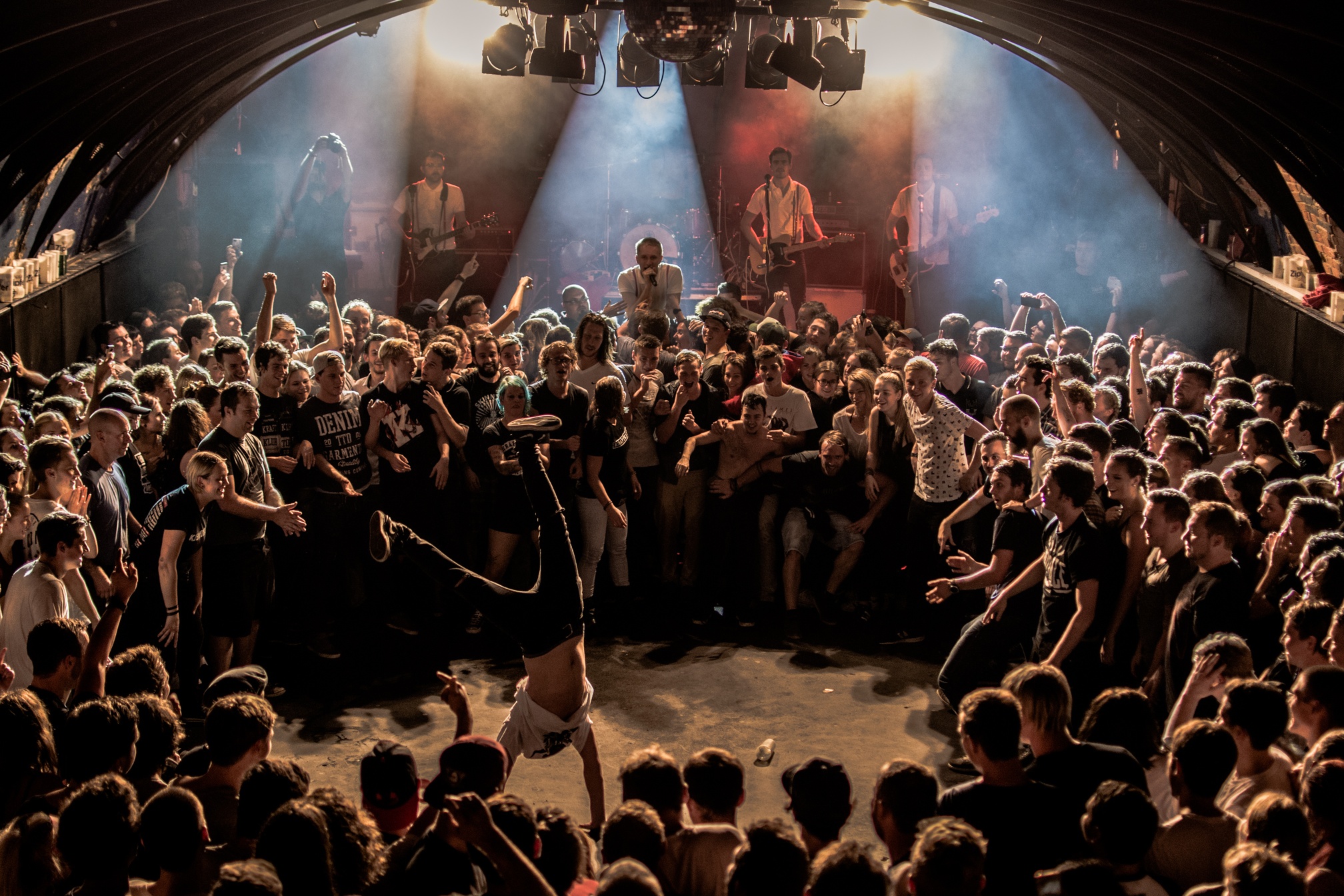 Eine Menschenmenge bei einem Konzert umringt eine Person, die auf ihren H&auml;nden Breakdance tanzt, w&auml;hrend auf der B&uuml;hne eine Band auftritt.