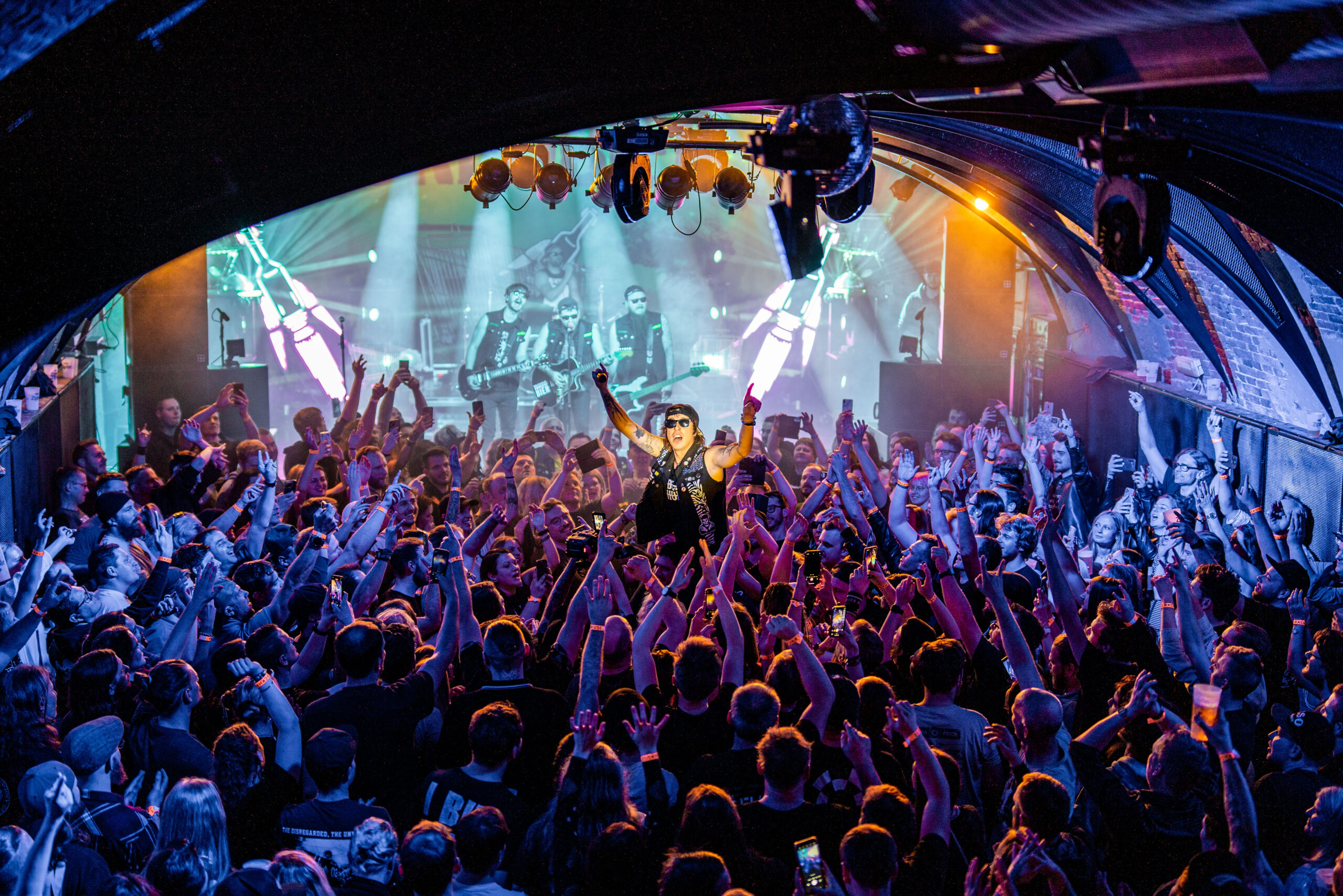 Eine Rockband tritt auf der B&uuml;hne eines &uuml;berf&uuml;llten, farbenfrohen Clubs auf, w&auml;hrend jubelnde Fans die H&auml;nde in die Luft recken.