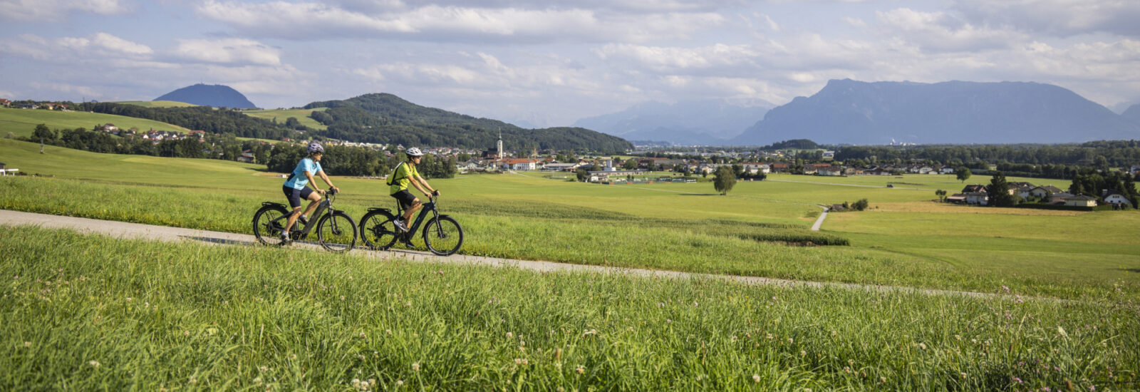 Zwei Menschen fahren mit dem Fahrrad auf einem Weg durch grüne Felder, im Hintergrund sind Hügel und Berge zu sehen, über ihnen ein bewölkter Himmel.
