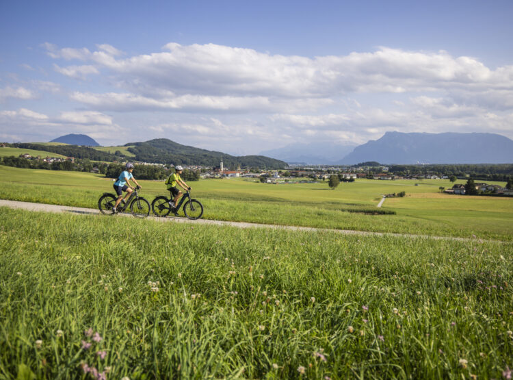 Zwei Menschen fahren mit dem Fahrrad auf einem Weg durch grüne Felder, im Hintergrund sind Hügel und Berge zu sehen, über ihnen ein bewölkter Himmel.