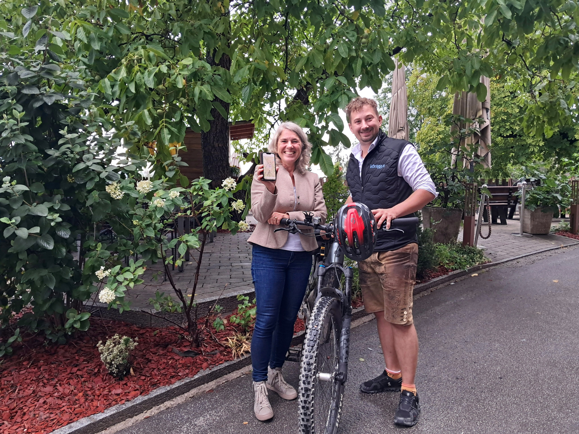 Zwei Menschen stehen draußen neben einem Fahrrad; einer hält ein Handy hoch, und beide lächeln unter grünen Bäumen.