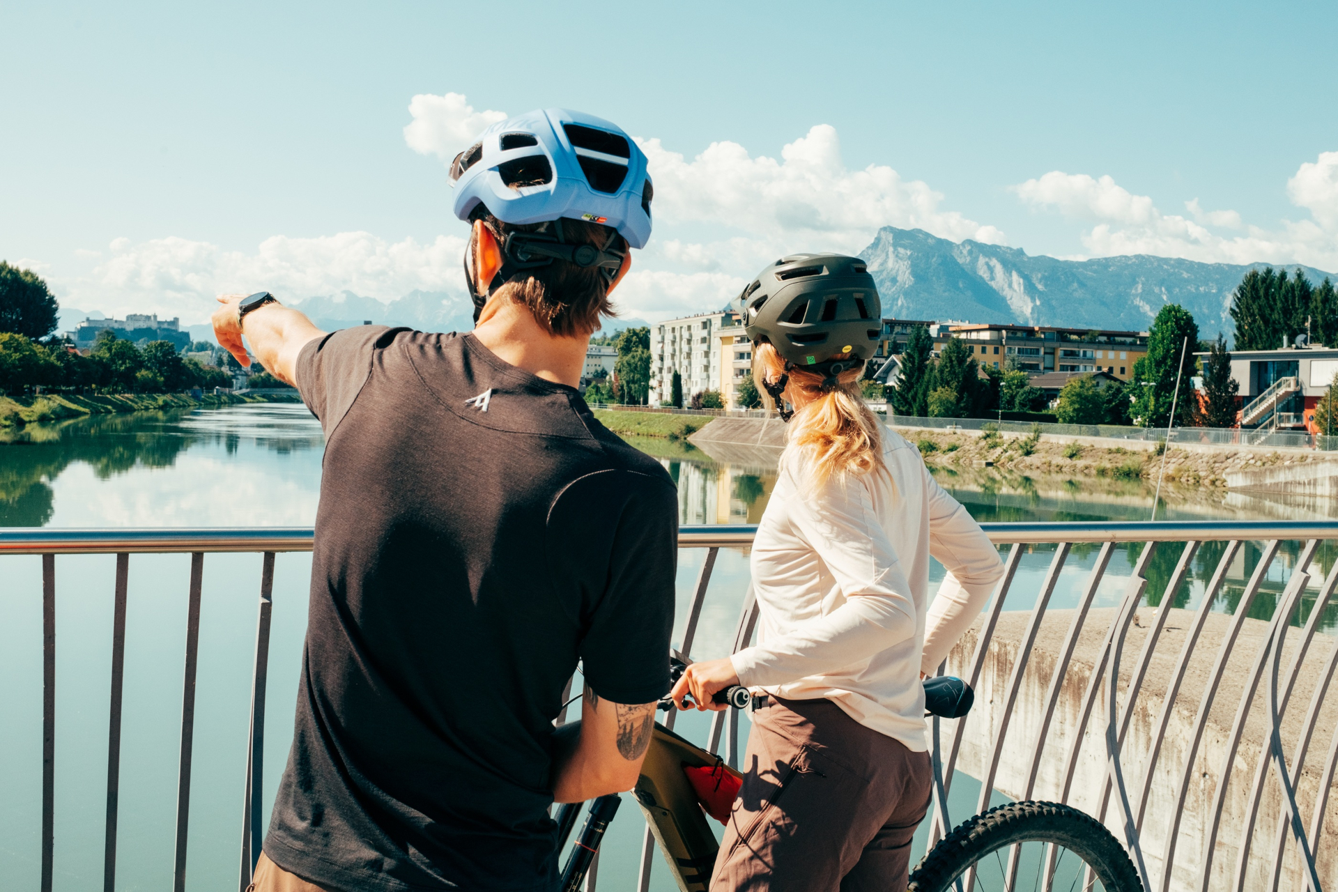 Zwei Radfahrer mit Helmen halten auf einer Brücke inne und blicken auf einen Fluss und die Berge in der Ferne.