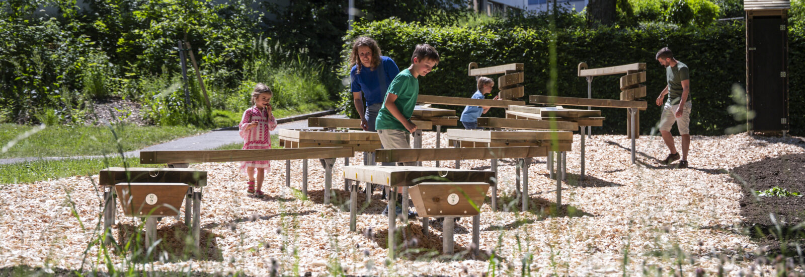 Kinder und Erwachsene spazieren um Holztische herum und spielen in einem sonnigen Park im Freien mit grünen Bäumen.