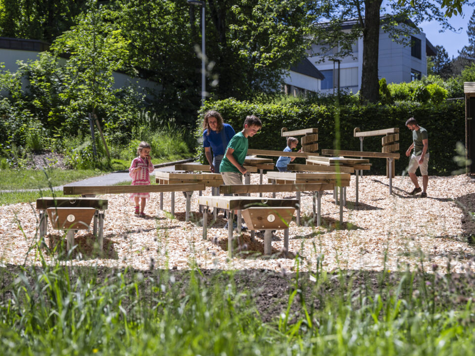 Kinder und Erwachsene spazieren um Holztische herum und spielen in einem sonnigen Park im Freien mit grünen Bäumen.