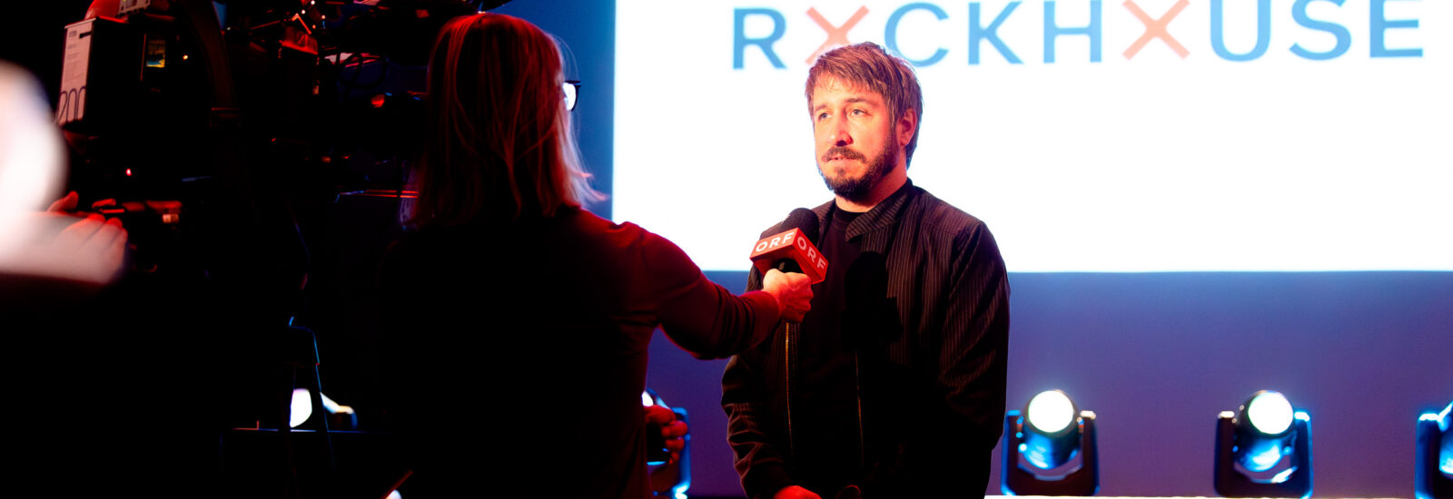 Eine Person wird auf der Bühne vor einem leuchtenden RYCKHOUSE-Schild und unter Bühnenlicht interviewt.