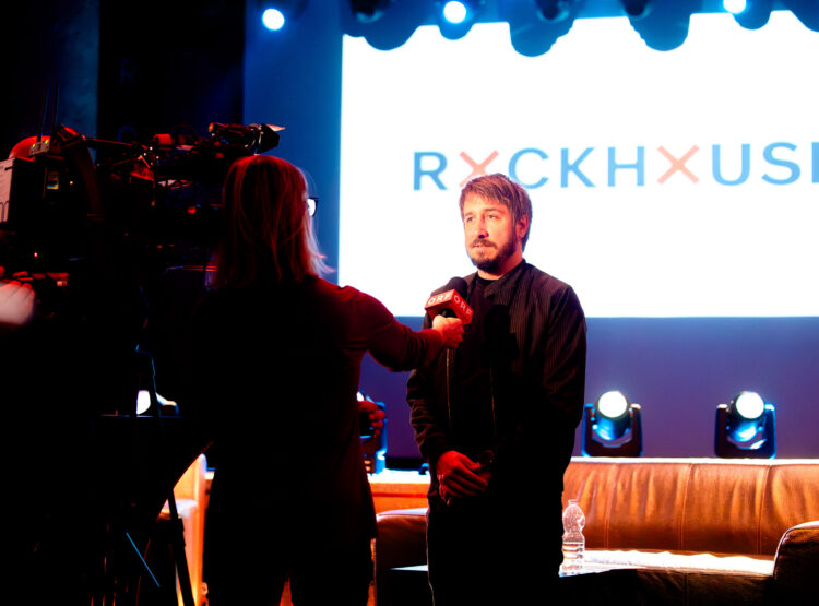 Eine Person wird auf der Bühne vor einem leuchtenden RYCKHOUSE-Schild und unter Bühnenlicht interviewt.