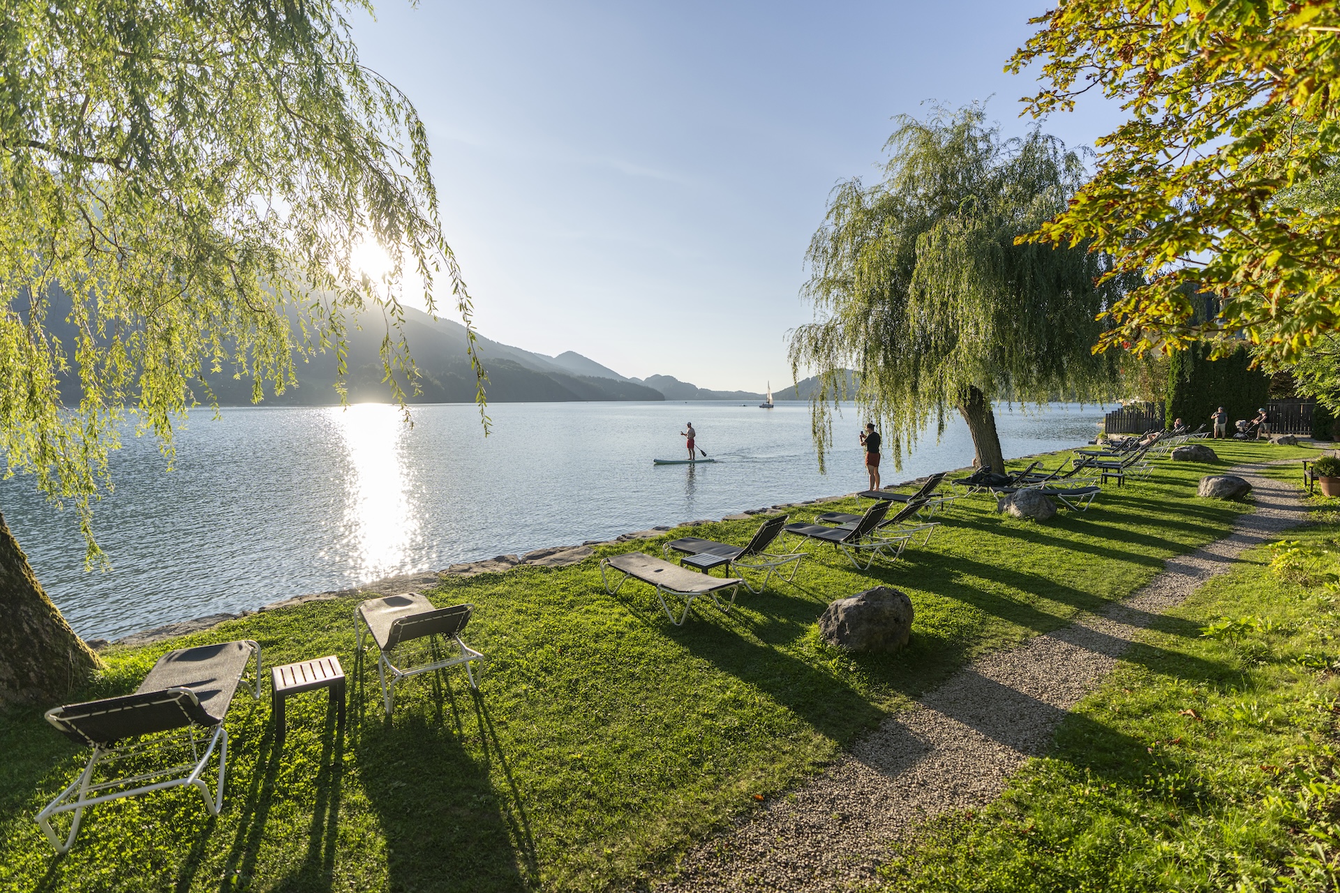 Eine Wiese am Seeufer mit leeren Liegestühlen, Bäumen und einer Person, die bei Sonnenuntergang auf dem Wasser Paddleboard fährt.