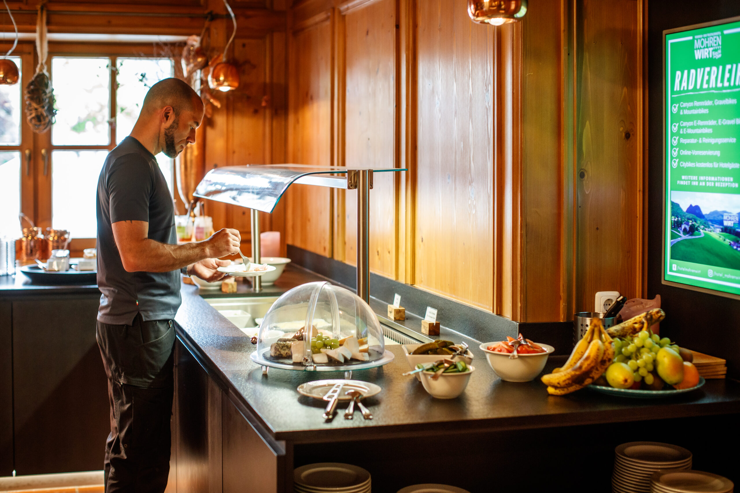 Ein Mann bedient sich an einem Buffet in einem holzgetäfelten Raum, in dem verschiedene Speisen und Obst angeboten werden.