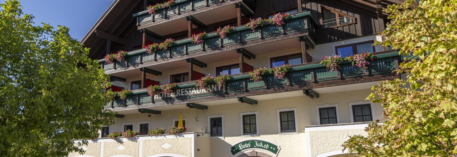 Ein sonniger Tag in einem traditionellen Hotel mit Balkonen, grünen Geländern und Blumen; davor stehen Bäume.