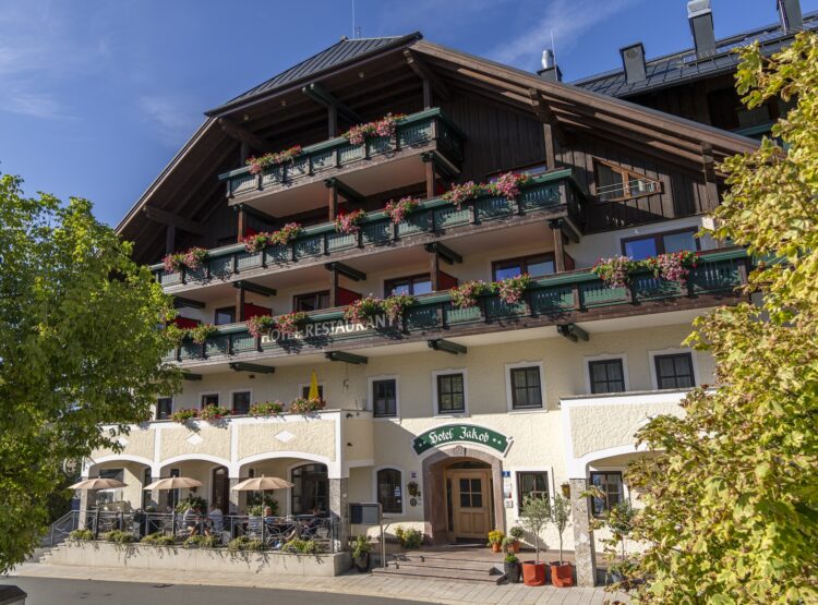 Ein sonniger Tag in einem traditionellen Hotel mit Balkonen, grünen Geländern und Blumen; davor stehen Bäume.