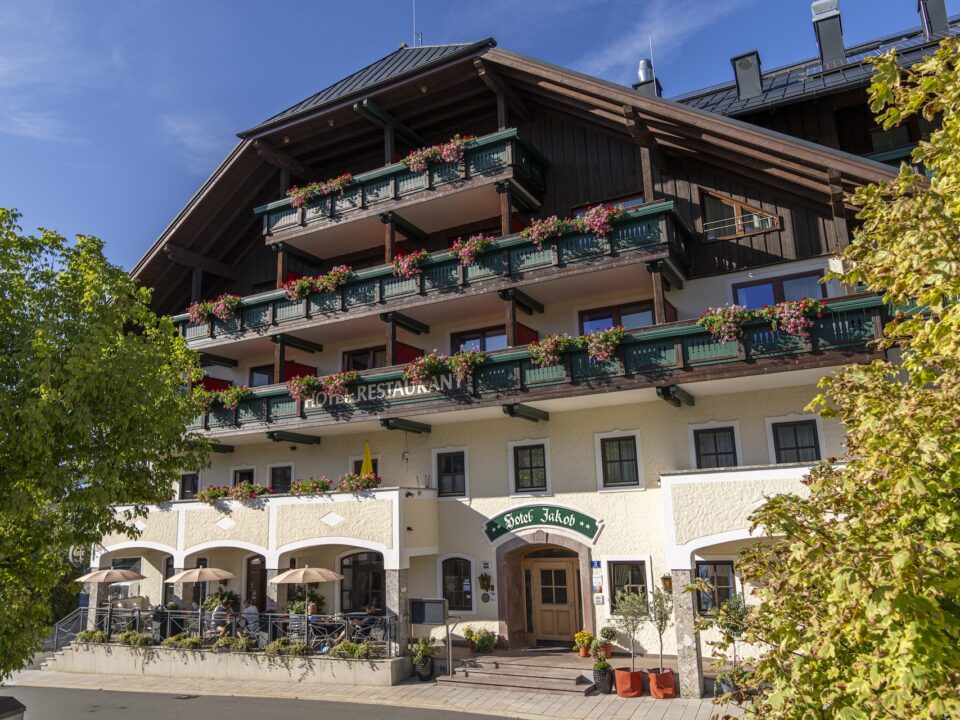 Ein sonniger Tag in einem traditionellen Hotel mit Balkonen, grünen Geländern und Blumen; davor stehen Bäume.
