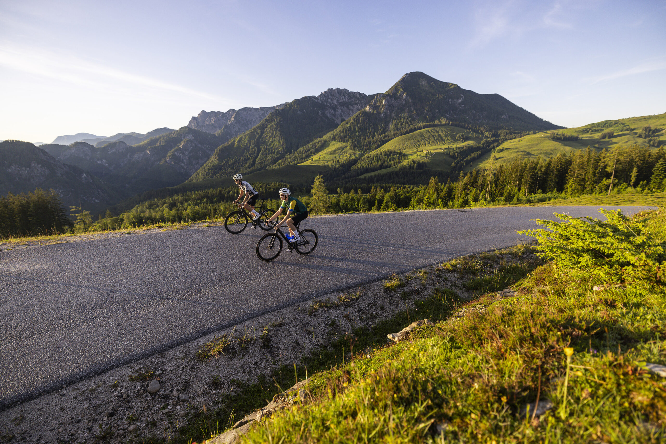 Zwei Menschen fahren mit dem Fahrrad auf einer Bergstraße, umgeben von grünen Hügeln und Bergen unter einem strahlend blauen Himmel.