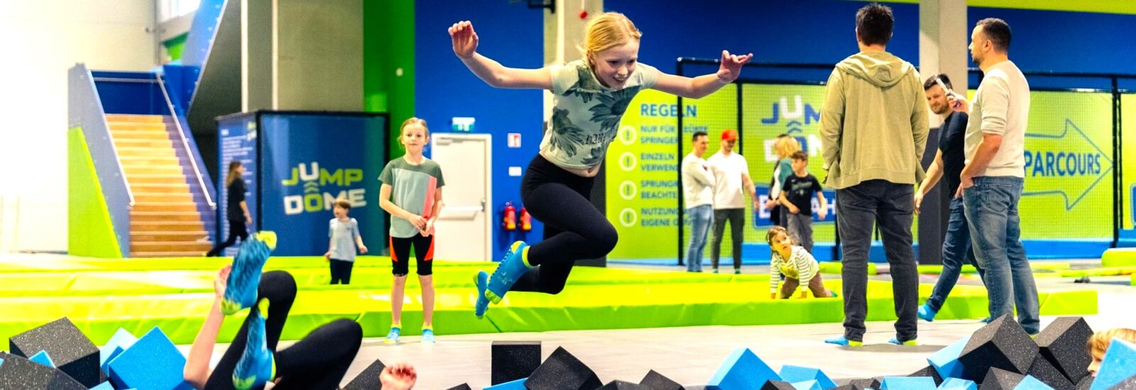 Eine Frau springt in einem Indoor-Trampolinpark in eine Schaumstoffgrube, während andere zuschauen und Kinder in der Nähe spielen.