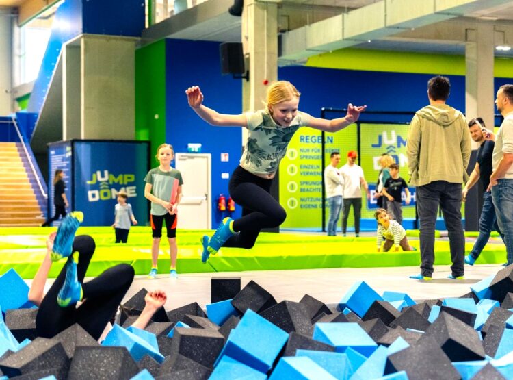 Eine Frau springt in einem Indoor-Trampolinpark in eine Schaumstoffgrube, während andere zuschauen und Kinder in der Nähe spielen.
