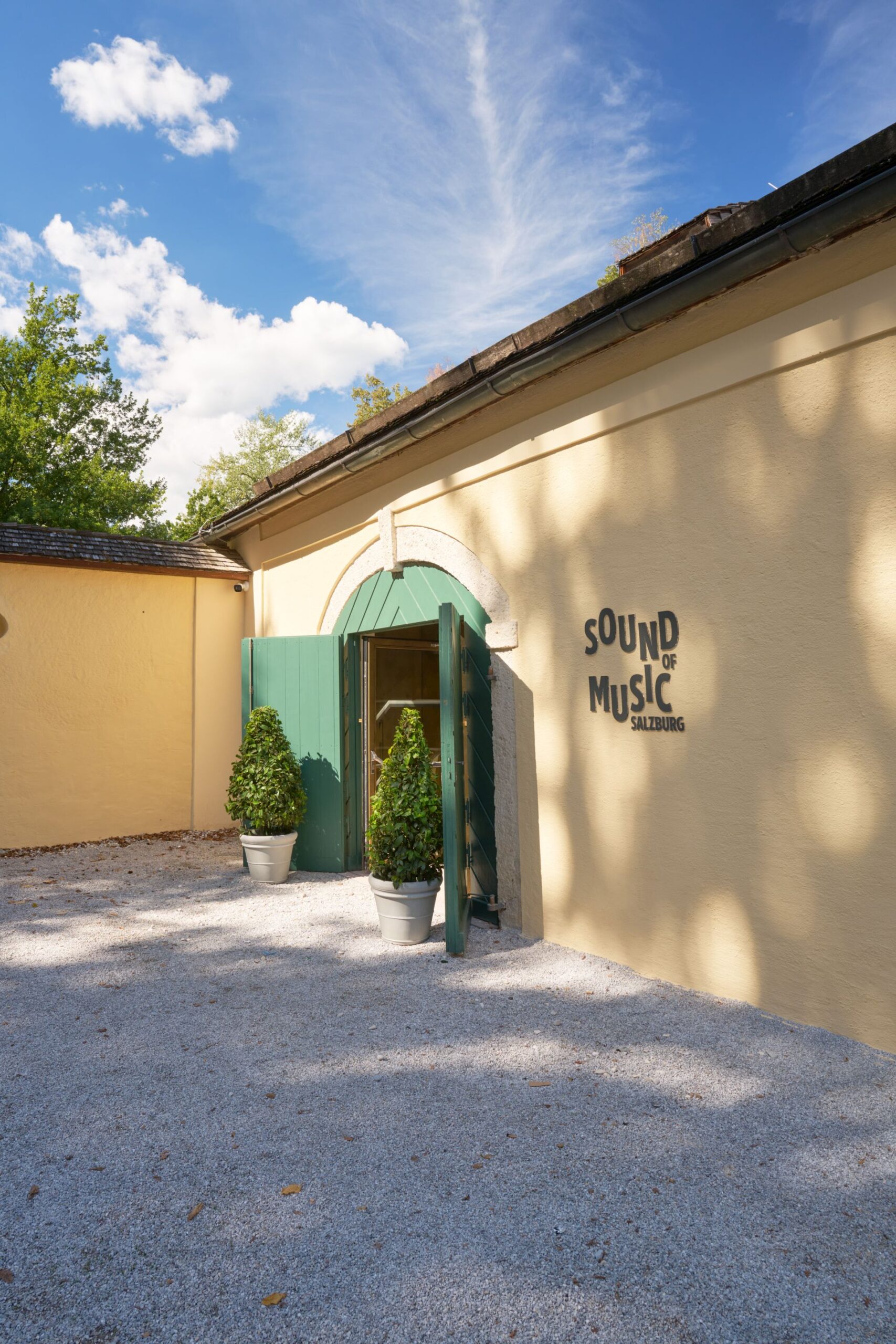 Gelbes Geb&auml;ude mit gr&uuml;ner T&uuml;r, zwei B&auml;umen in T&ouml;pfen und der Aufschrift &bdquo;Sound of Music Salzburg&ldquo; an der Wand.