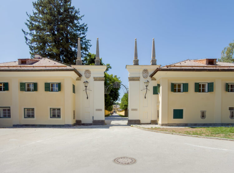 Zwei cremefarbene Gebäude mit grünen Fensterläden und spitzen Säulen, die durch eine breite Zufahrt voneinander getrennt sind.