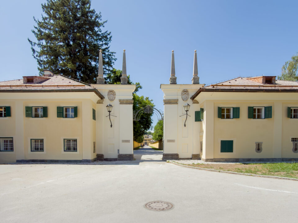 Zwei cremefarbene Gebäude mit grünen Fensterläden und spitzen Säulen, die durch eine breite Zufahrt voneinander getrennt sind.