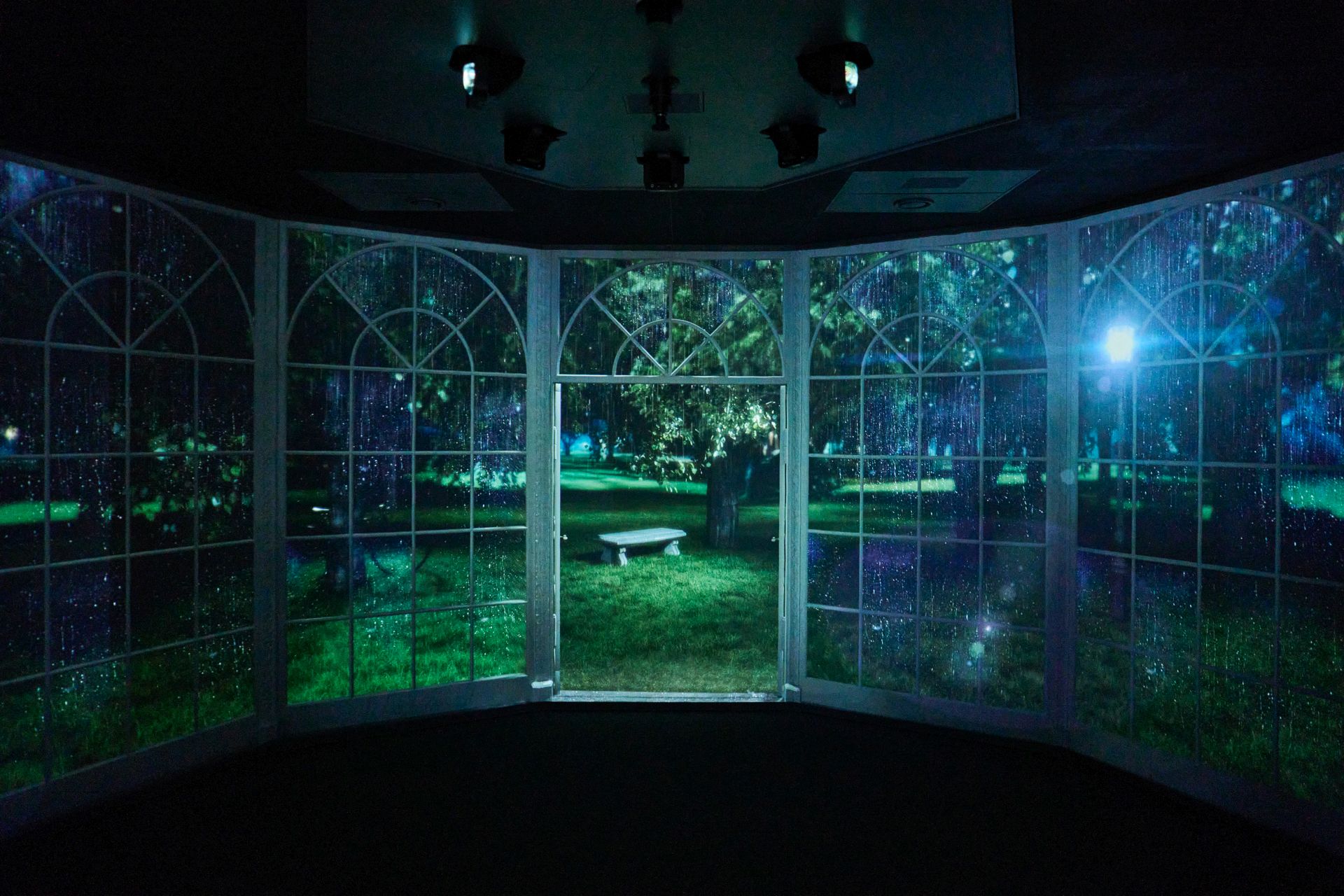 Große Fenster geben den Blick auf einen nächtlichen Garten mit einer Bank frei, der durch die gewölbten Scheiben in blaues Licht getaucht ist.