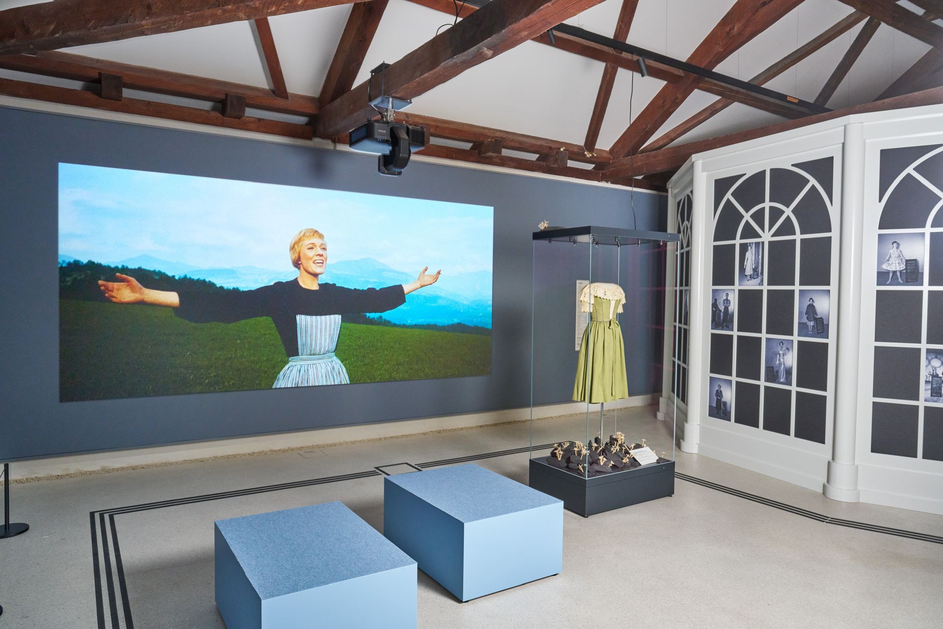 Ausstellungsraum mit einer gro&szlig;en Leinwand, auf der eine Frau auf einem Feld zu sehen ist, sowie ausgestellten Kost&uuml;men und Fotos.