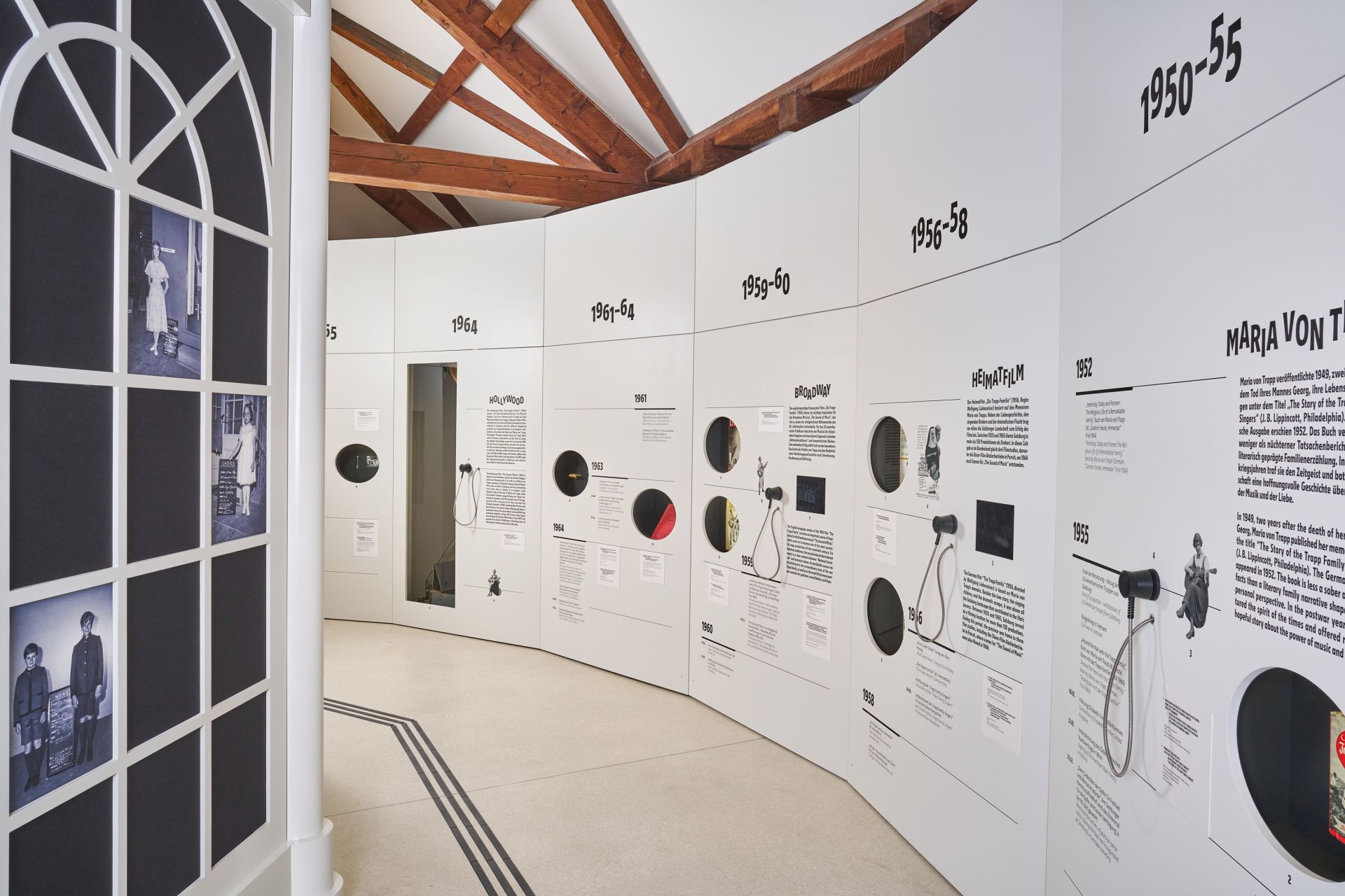 An einer weißen, geschwungenen Museumswand sind Exponate und Texte nach Jahrzehnten von 1950 bis 1968 ausgestellt; Foto im Fenster links.