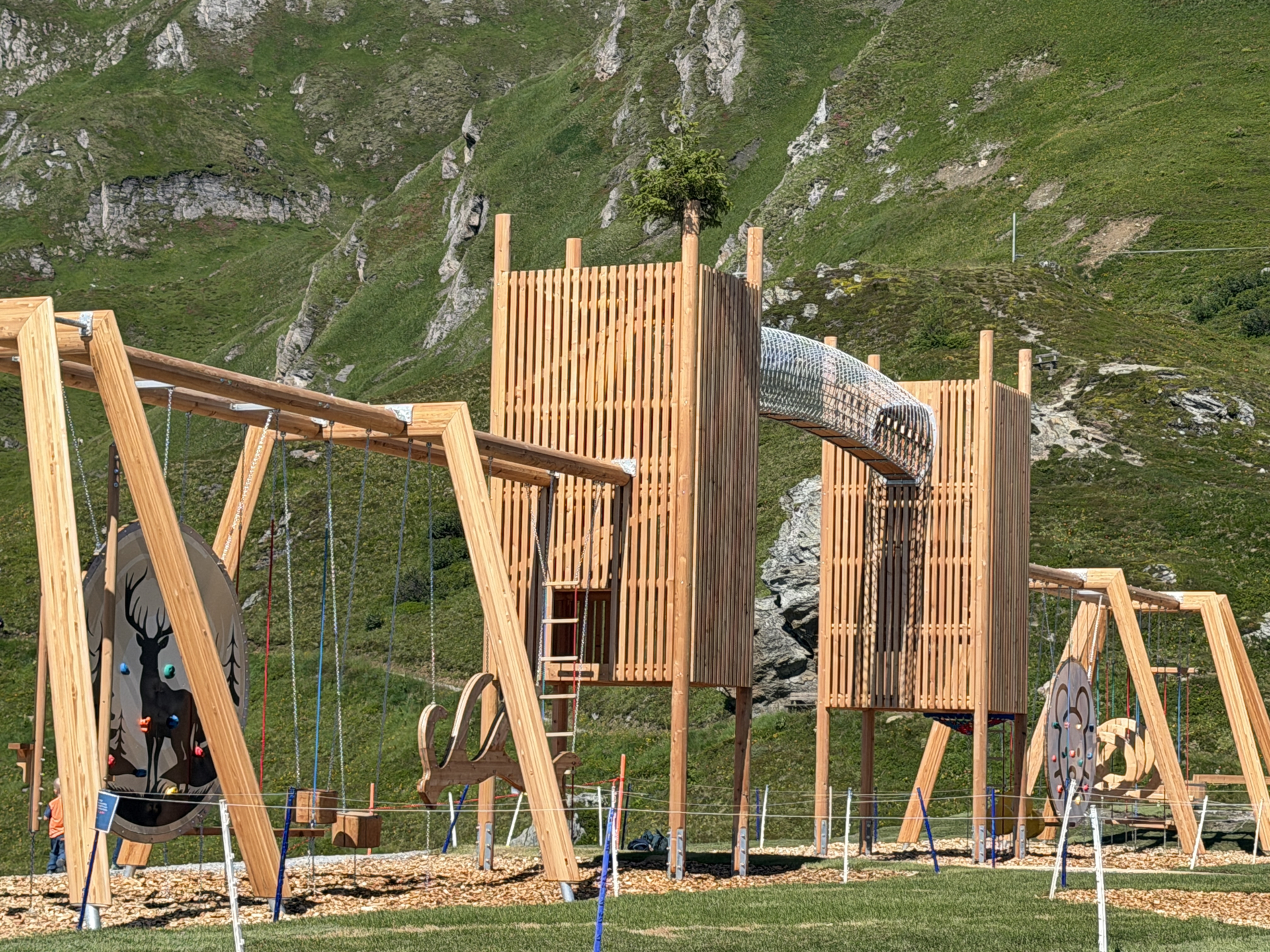 Holzspielplatz mit Schaukeln und Rutschen vor der Kulisse eines grasbewachsenen Berges an einem sonnigen Tag.