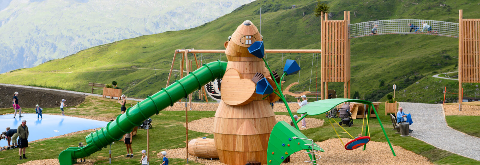 Kinder spielen auf einem Spielplatz aus Holz mit Rutschen, Schaukeln und einer Kletterwand in einer bergigen, grasbewachsenen Landschaft.