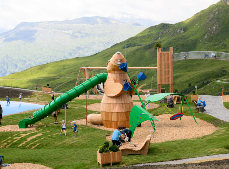Kinder spielen auf einem Spielplatz aus Holz mit Rutschen, Schaukeln und einer Kletterwand in einer bergigen, grasbewachsenen Landschaft.