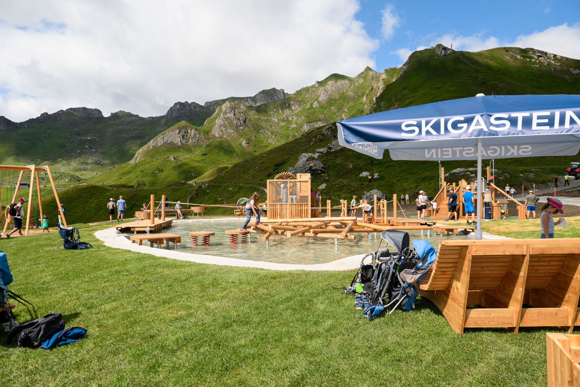 Menschen spielen in einem Wasser-Erlebnispark aus Holz inmitten grasbewachsener Berge, &uuml;ber dem ein Sonnenschirm mit der Aufschrift &bdquo;SKIGASTEIN&ldquo; steht.