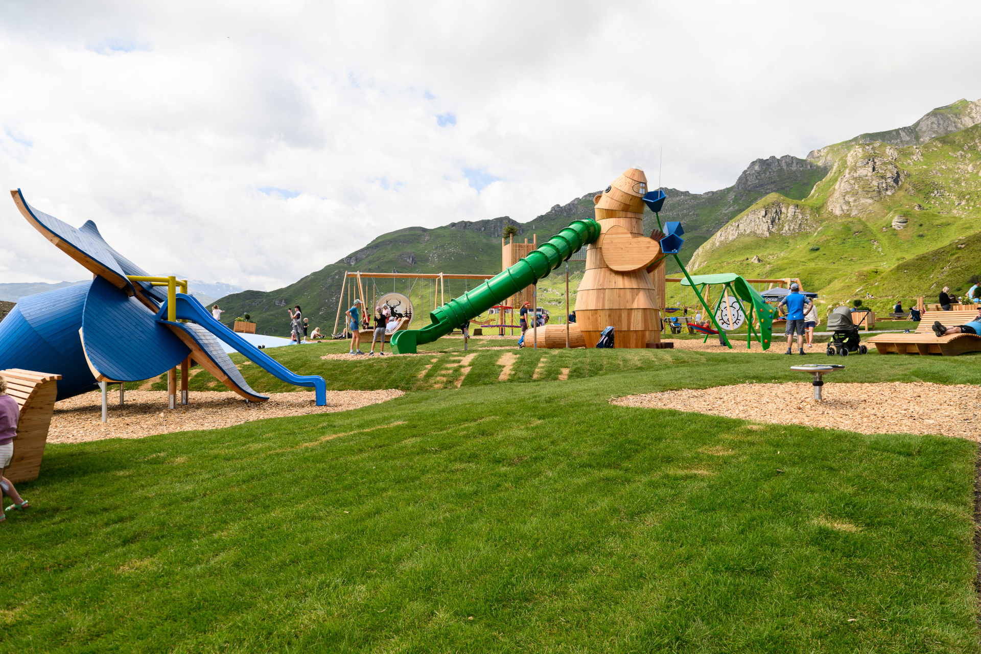 Kinder spielen auf Spielger&auml;ten aus Holz und in Blaut&ouml;nen, im Hintergrund sind grasbewachsene H&uuml;gel und Berge zu sehen.