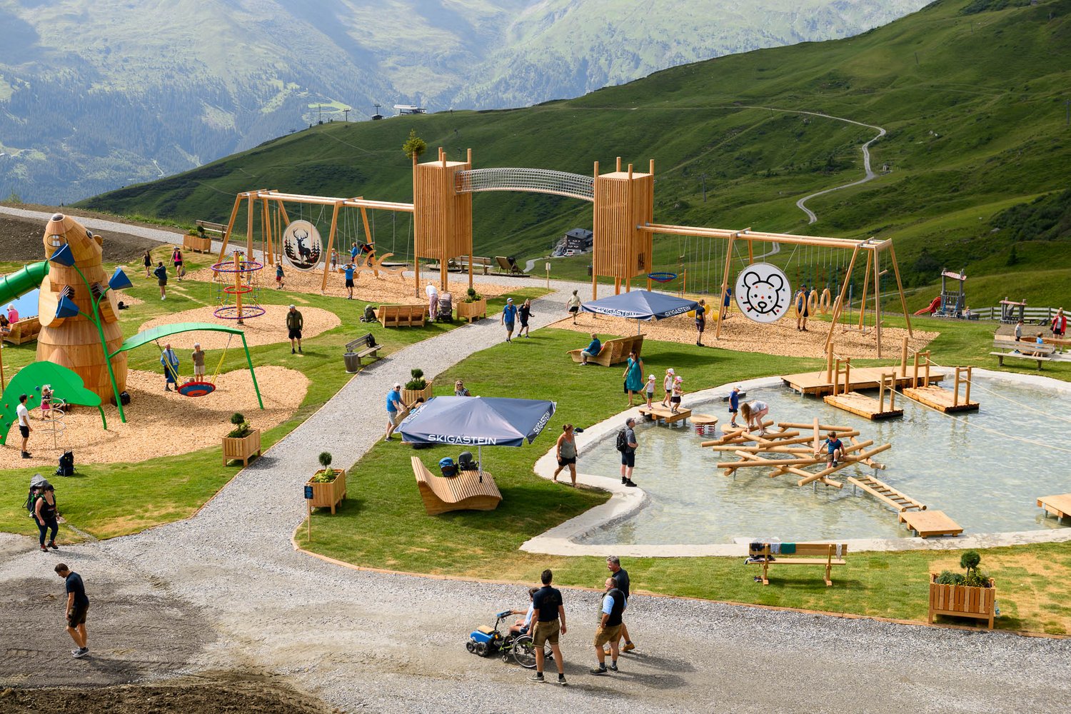 Kinder und Erwachsene spielen auf einem Holzspielplatz und in einem flachen Teich in einer bergigen, grasbewachsenen Gegend.