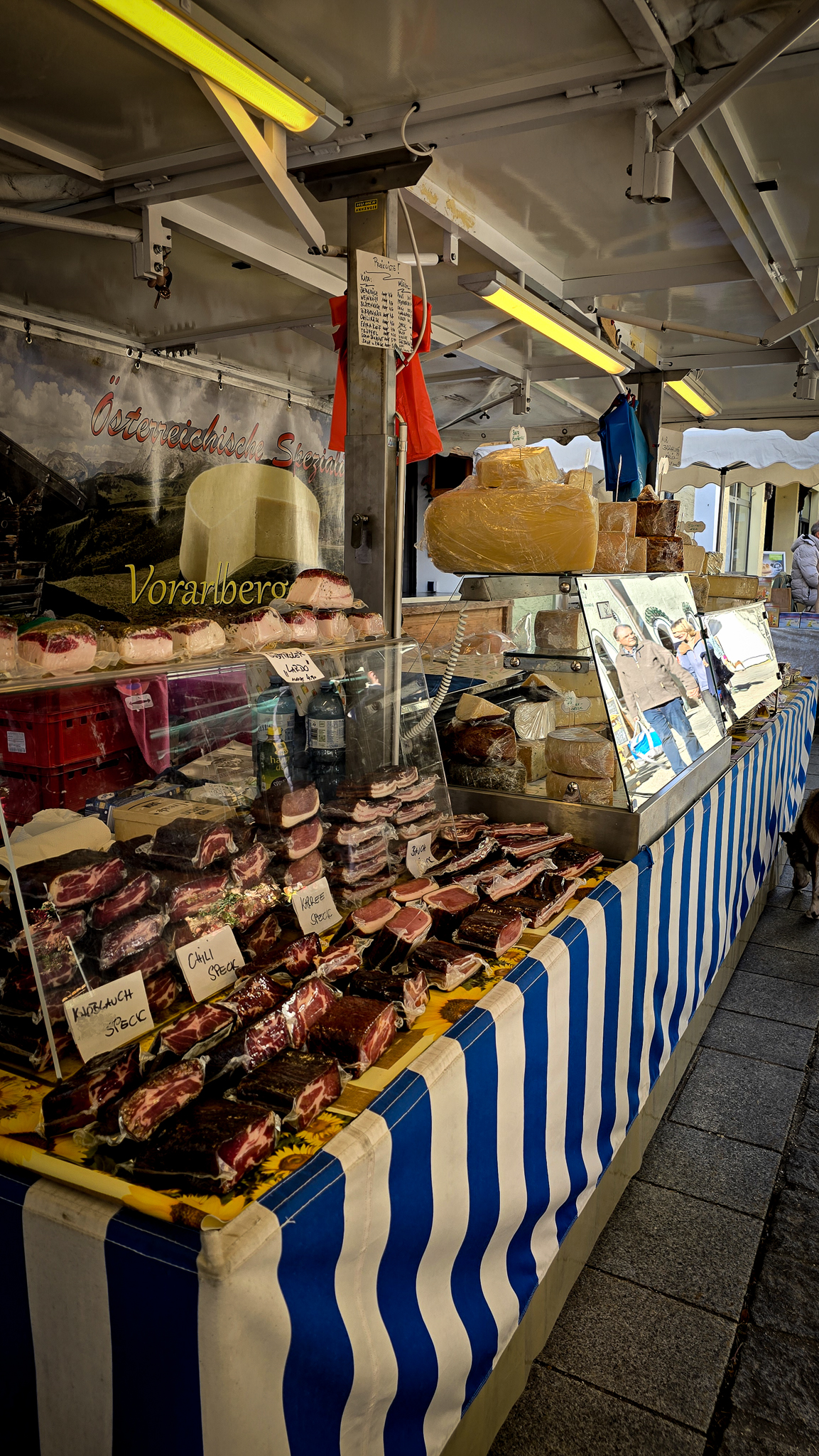 Marktstand auf dem Wolfgangi-Kirtag mit K&auml;se, Wurstwaren und einer blau gestreiften Tischdecke, Seitenansicht.