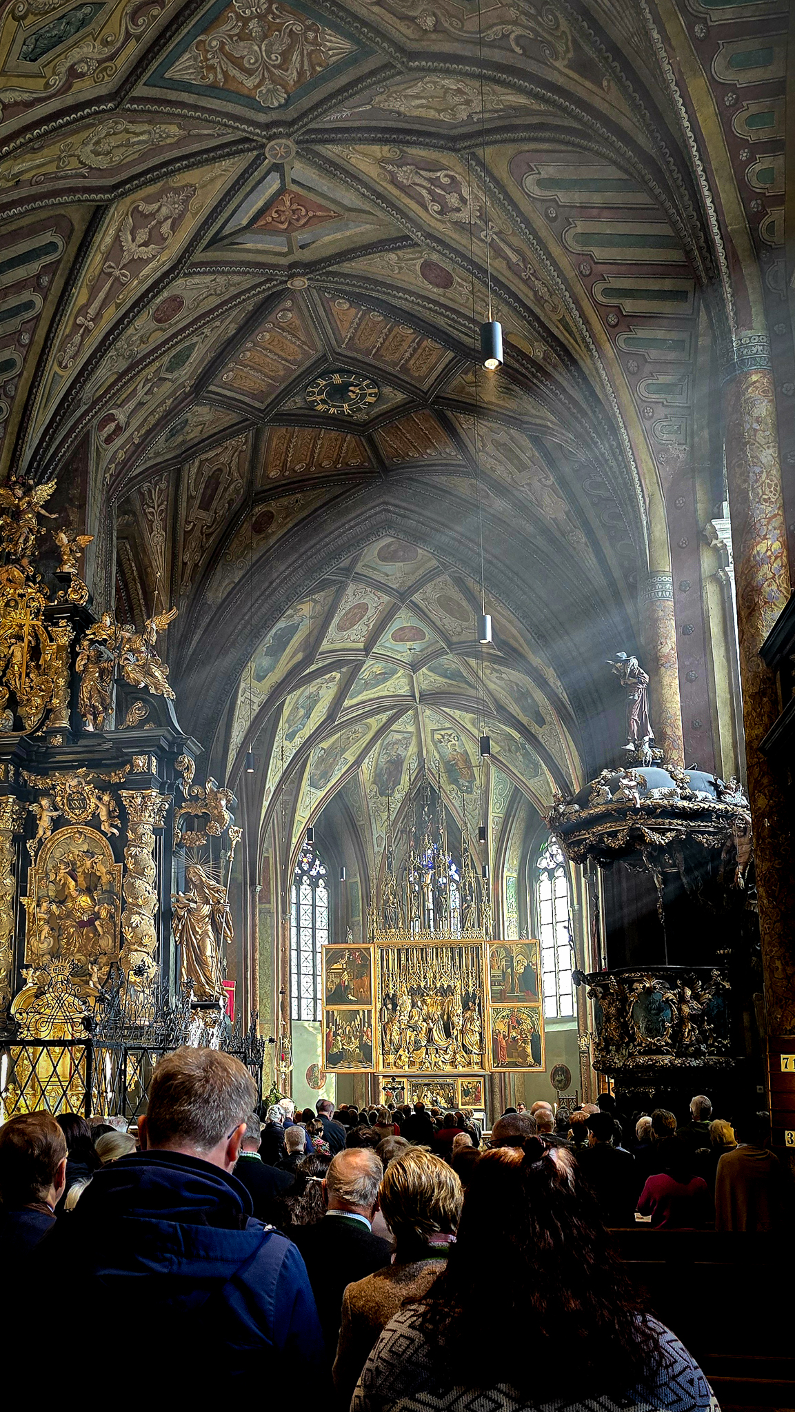 Menschen sitzen w&auml;hrend des Wolfgangi-Kirtags in einer pr&auml;chtigen, reich verzierten Kirche, w&auml;hrend Sonnenlicht durch die Buntglasfenster f&auml;llt.