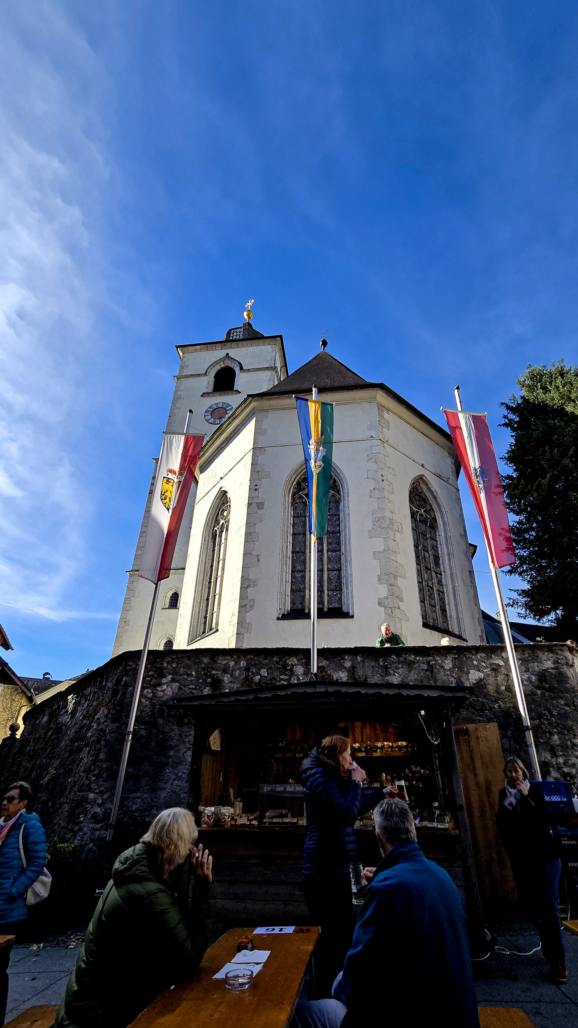 W&auml;hrend des Wolfgangi-Kirtags sitzen Menschen an Holztischen vor einer wei&szlig;en Kirche, &uuml;ber ihnen strahlt ein strahlend blauer Himmel.