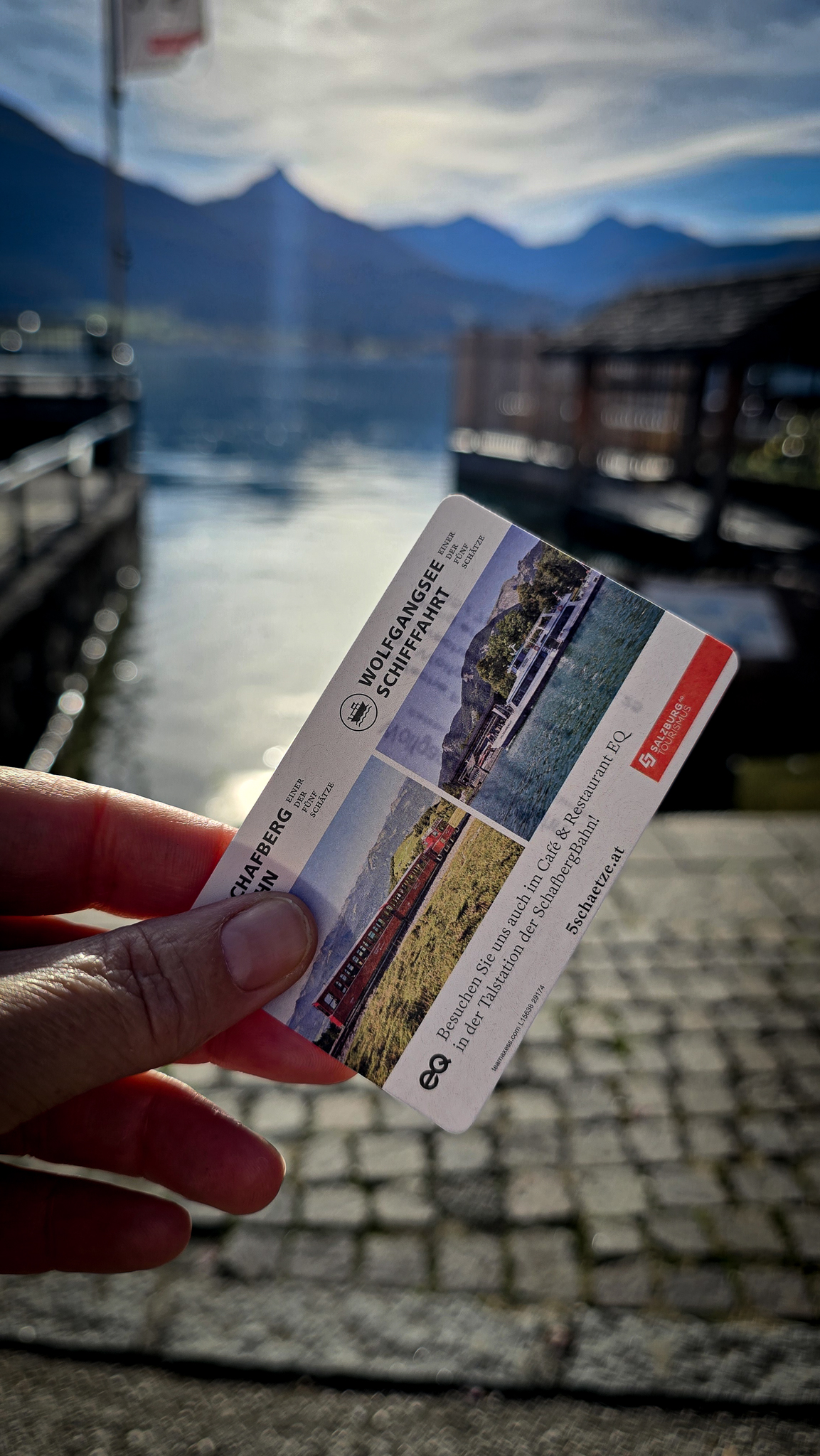 Eine Hand h&auml;lt am See w&auml;hrend des Wolfgangi-Kirtags eine Fahrkarte der Wolfgangsee-Schifffahrt in der Hand, im Hintergrund sind Boote und Berge zu sehen.