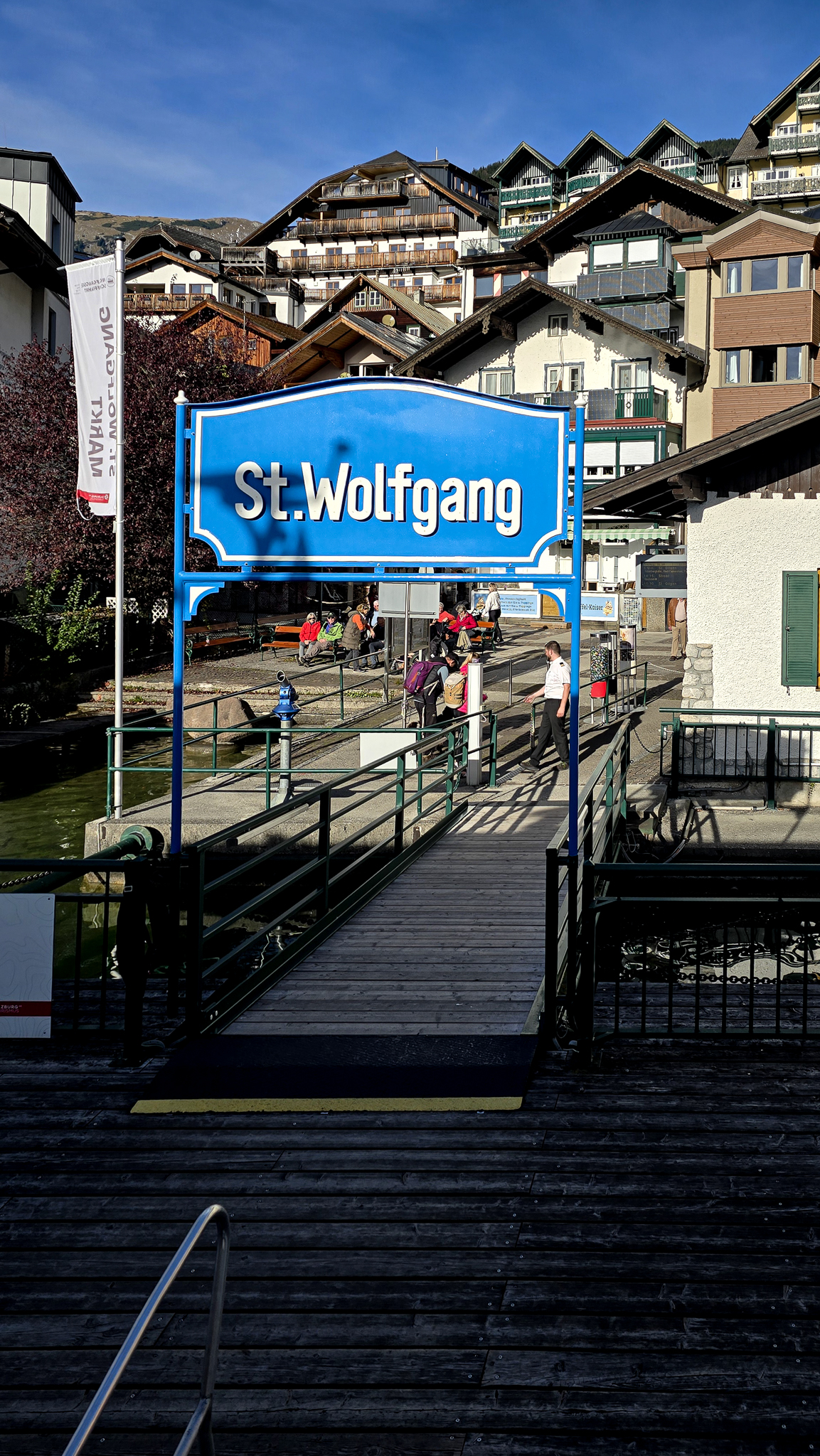 Ein blaues &bdquo;St. Wolfgang&ldquo;-Schild an einem Steg, im Hintergrund Menschen und die Feierlichkeiten zum &ouml;sterreichischen Wolfgangi-Kirtag.
