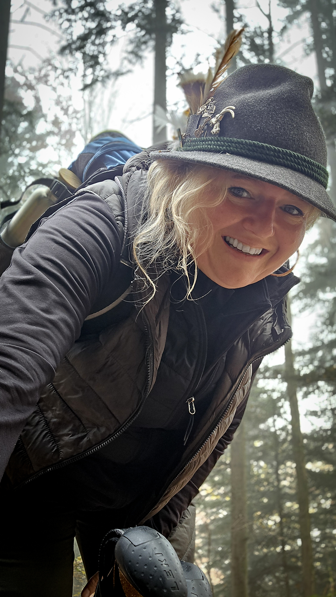 Eine l&auml;chelnde Person mit einem Federhut und einem Rucksack, die durch einen nebligen Wald wandert und bereit ist f&uuml;r das Wolfgangi-Kirtag-Abenteuer.