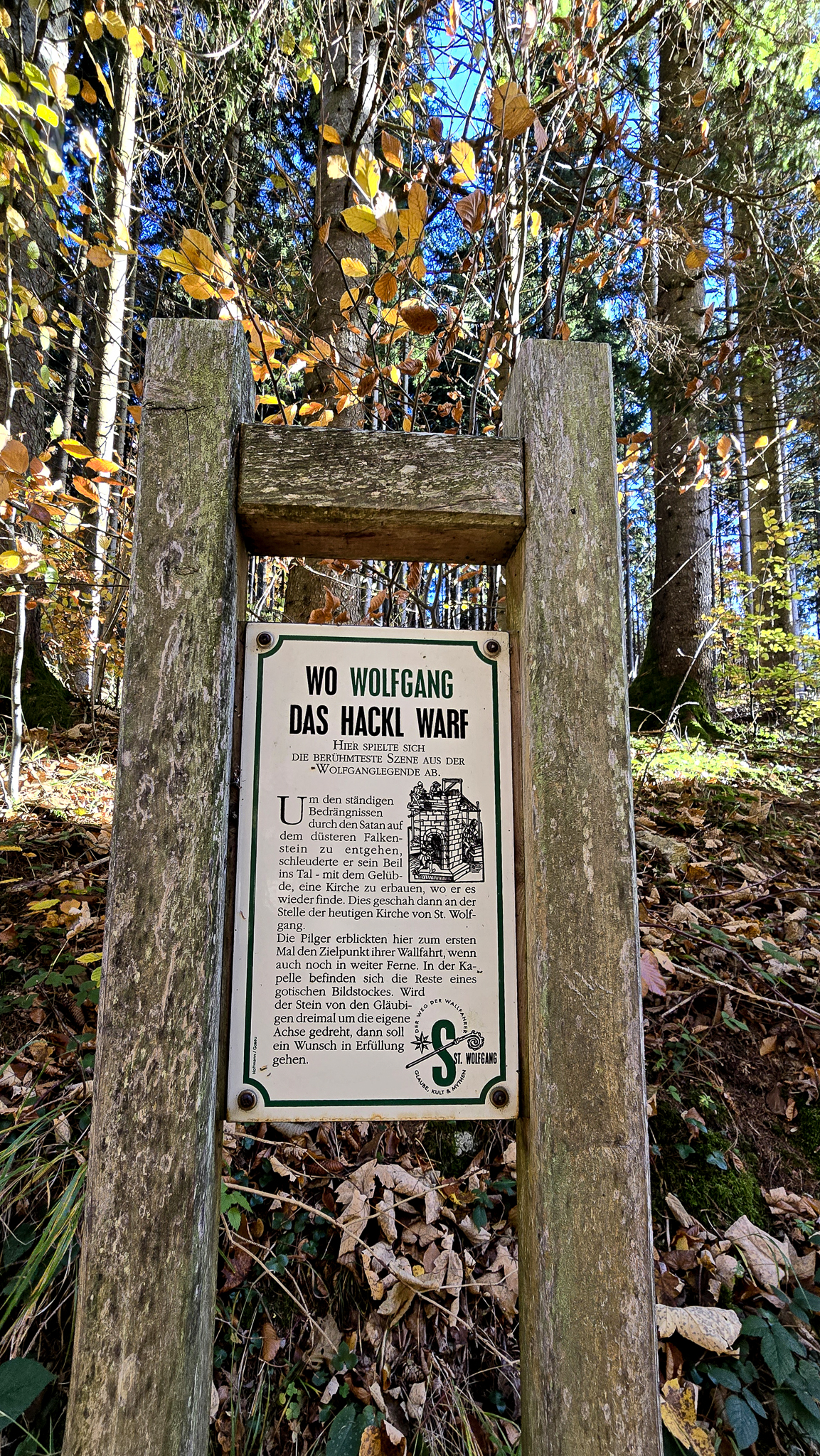 Ein Schild mit Holzrahmen f&uuml;r den Wolfgangi-Kirtag und deutscher Aufschrift steht in einem Wald mit hohen Herbstb&auml;umen.