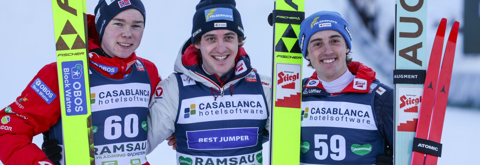 Drei Skispringer posieren gemeinsam auf einem Podium, halten ihre Skier in den Händen und tragen Sportbekleidung mit Sponsorenlogos.