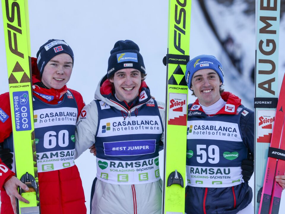 Drei Skispringer posieren gemeinsam auf einem Podium, halten ihre Skier in den Händen und tragen Sportbekleidung mit Sponsorenlogos.